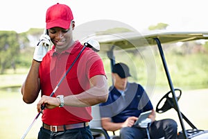 Checking in with home base. a young man having a conversation on his phone on a golf course while his friend sits in the