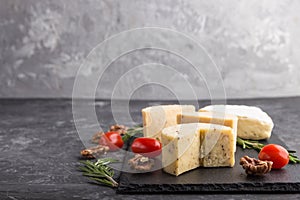 Cheddar cheese and various types of cheese with rosemary and tomatoes on black slate board on a black concrete background. Side