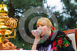 Cheeked Russian cheerful young woman with a scarf draped pavloposadskie, sitting at a table with a samovar and drinking tea with photo