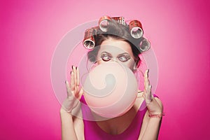 Cheeky girl with bubble gum posing on pink background in body, with curlers on head. Pretty woman with sweet makeup making