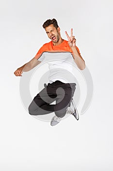 Cheeky, happy attractive young bearded masculine sportsman in activewear, jumping and showing peace sign in air, wink