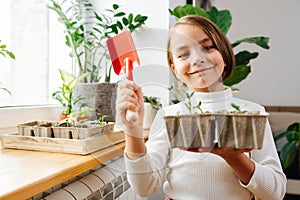 Cheeky little girl holding seedlings and a small red shovel, next to a window