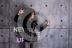 Cheer up. Cheerful young curly woman dancing on concrete wall background. Multi-colored projection says: Never give up.