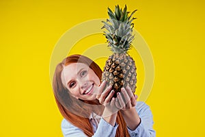 Cheerfu redhaired ginger woman hiding face behind pineapple on yellow color background