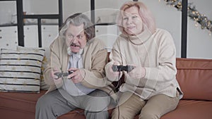 Cheerful absorbed senior man and woman playing video games with game consoles. Portrait of joyful old couple having fun