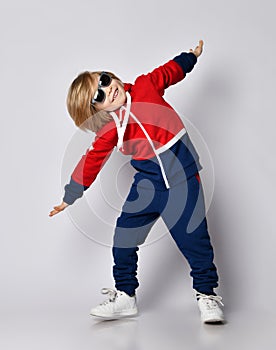 Cheerful active frolic blond kid boy in blue and red hoodie, pants and sunglasses is playing aircraft pilot