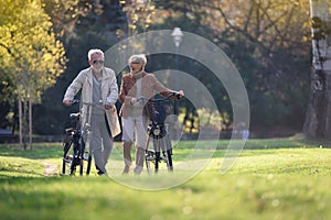 Cheerful active senior couple with bicycles walking through park together