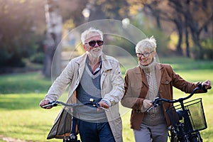 Cheerful active senior couple with bicycles walking through park together