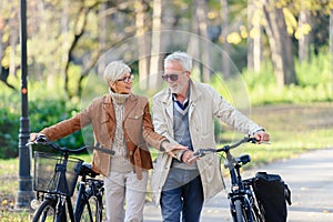 Cheerful active senior couple with bicycles walking through park together