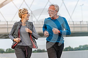 Cheerful active senior couple jogging together outdoors along the river