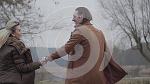 Cheerful adult Caucasian couple running outdoors holding hands. Joyful brunette man hugging and spinning blond smiling