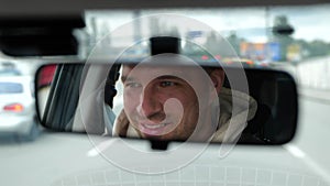 Cheerful adult Caucasian male driving a car in city looking and smiling in rearview mirror. Happy man face reflected in