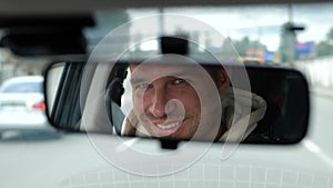 Cheerful adult Caucasian male driving a car in city looking and smiling in rearview mirror. Happy man face reflected in