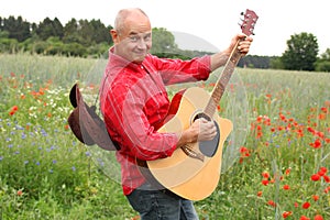 Cheerful adult man in a red shirt and a coboy hat on a field of green poppies plays guitar, posing and having fun, concept musical
