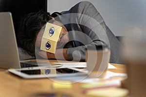 Cheerful adult man resting in office with sticky notes on the eyes. Sleeping man having stickers with eyes painted on face