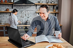 Cheerful adult man sit at table in kitchen. He look on laptop and touch it. Guy hold cup of coffee. His wife stand at