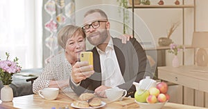 Cheerful adult son and mature mother making video call with smartphone while sitting at home. Elderly woman and young