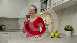 Cheerful adult woman eating bar of chocolate, savoring dessert at home