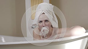 Cheerful adult woman taking bath foam and blowing it to camera. Portrait of happy young Caucasian lady in hair towel