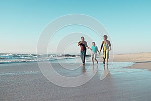 Cheerful african american boy holding father and mother's hands and running in sea against sky