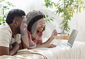 Cheerful black couple using laptop, lying on bed