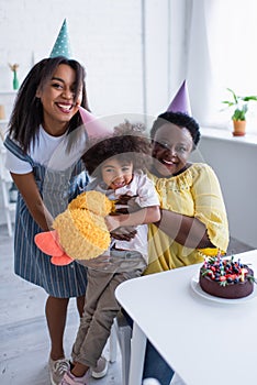 cheerful african american girl holding soft toy while mom and granny hugging her near birthday cake,stock image