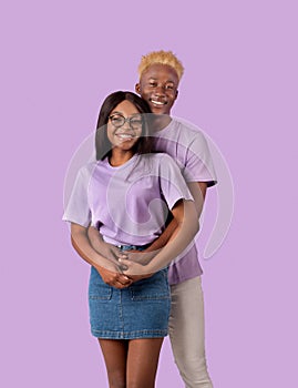 Cheerful African American guy hugging his beloved woman from behind, smiling and looking at camera on lilac background