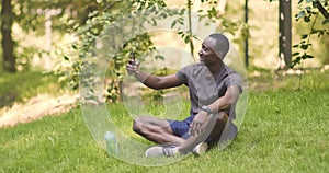 Cheerful african american guy talking on mobile phone, sitting on grass in park