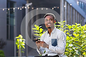 Cheerful african american guy using mobile phone sitting on bench in city park outdoors. Happy black man in casual clothes with a