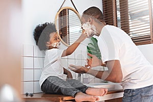 Cheerful African American Man and little boy having fun laughing with shaving foam on their faces in bathroom at home together