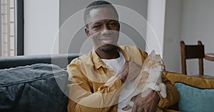 Cheerful African American man stroking cat petting domestic animal expressing love to cat on sofa at home