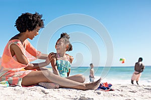 Cheerful african american mother and daughter sitting together while father and son playing at beach