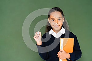 cheerful african american schoolgirl holding piece of chalk and book while standing near chalkboard.