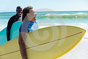 Cheerful african american senior couple carrying surfboards walking at beach on sunny day