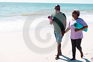Cheerful african american senior couple with mats holding hands while walking at beach on sunny day