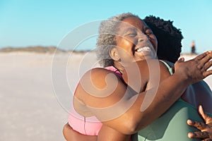 Cheerful african american senior mother embracing young daughter while enjoying at beach against sky