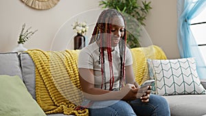 Cheerful african american woman with braids enjoying home comforts, sitting on sofa and having fun texting online using her