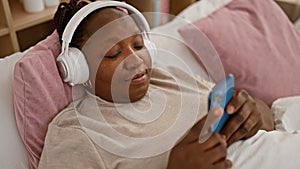 Cheerful african american woman chilling at home, happily texting and jamming to her favorite tunes while lying in bed using her