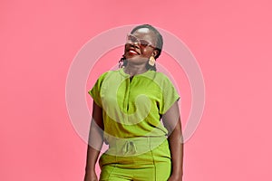 Cheerful African-American woman dressed vibrant attire looking at camera with smile against pastel pink studio