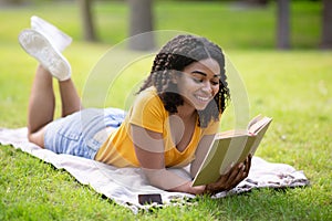 Cheerful African American woman reading interesting book while lying on green lawn at park