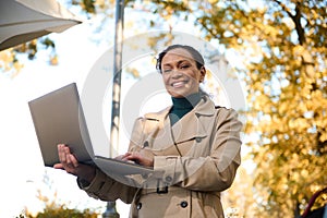 Cheerful African American woman using laptop, typing on keyboard on the autumnal park. Distant work concept, outdoor business.