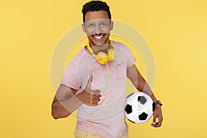 Cheerful african american young man holding soccer ball showing thumb up over yellow background.