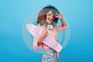 Cheerful african female model in jeans posing with peace sign holding longboard in studio. Laughing mulatto girl wears