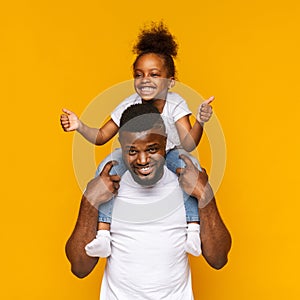 Cheerful african girl gesturing thumbs up, sitting on daddy`s shoulders