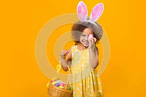 A cheerful african girl with rabbit ears on her head with a basket of colored eggs in her hands