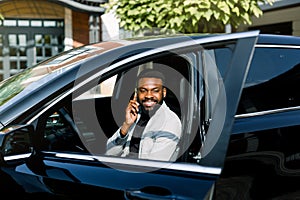 Cheerful African man in gey jacket sitting in car talking by the phone, office center on the background