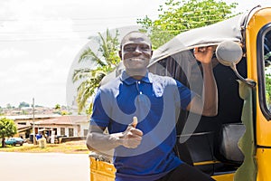 cheerful african man standing next to his tuk tuk taxi smiling and giving a thumbs up