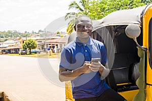 cheerful african man standing next to his tuk tuk taxi smiling and using his smart phone