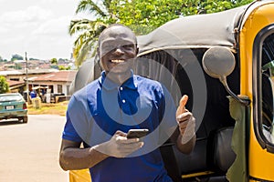 cheerful african man standing next to his tuk tuk taxi smiling and using his smart phone giving a thumbs up