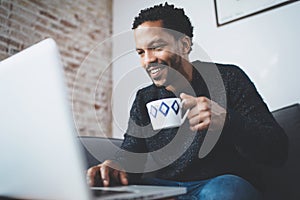 Cheerful African man using computer and smiling while sitting on the sofa.Black guy holding ceramic cup in hand.Concept